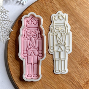 May include: Two plastic cookie cutters shaped like a nutcracker soldier. One is pink, the other white. Both feature a crown, moustache, and uniform details. Perfect for festive baking.