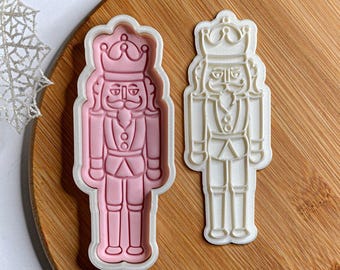 May include: Two plastic cookie cutters shaped like a nutcracker soldier. One is pink, the other white. Both feature a crown, moustache, and uniform details. Perfect for festive baking.