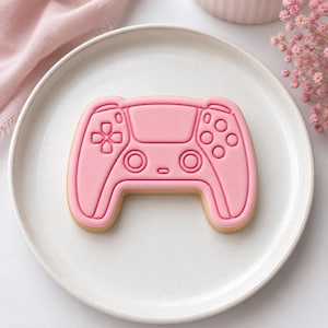 Peut inclure: Un biscuit rose clair en forme de manette de jeu vidéo avec des détails en relief, notamment une croix directionnelle, deux joysticks circulaires et quatre boutons d'action. Le biscuit est centré sur une assiette blanche au fini lisse.