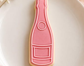 May include: A champagne bottle shaped sugar cookie decorated with smooth pink icing. The cookie features raised piped icing details outlining the bottle neck, a circular emblem on the body, and a rectangular label area at the base of the bottle.