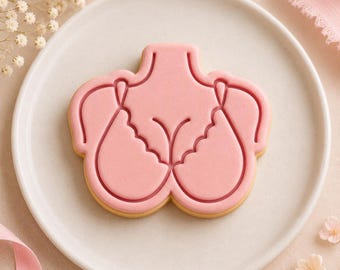 May include: A light pink cookie shaped like a torso wearing a bra, featuring embossed lines that define the neckline, straps, and scalloped edges of the garment. The cookie has a smooth, matte finish and is centered on a round, off-white plate.