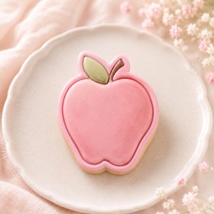 Könnte beinhalten: Ein Keks in Apfelform mit glattem rosa Zuckerguss, einem kleinen grünen Blatt und einem braunen Stiel. Der Keks liegt mittig auf einem runden, cremefarbenen Teller mit gewelltem Rand und zeigt eine glatte Oberfläche sowie klare Konturen.