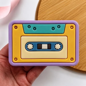 May include: A rectangular silicone mold shaped like a vintage cassette tape. The frame is lavender, featuring a yellow faceplate, a teal top section, a cream-colored center, and a blue horizontal strip with two white circular gear details.