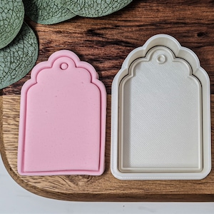 Könnte beinhalten: Ein weißer Plastik-Ausstecher in Form eines Etiketts mit einem gewellten Rand. Der Ausstecher wird neben einem rosa Silikon-Ausstecher der gleichen Form gezeigt.
