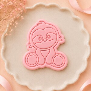 May include: A light pink cookie cutter shaped like a cartoon sloth with large eyes, a small nose, and rounded feet. The design features embossed lines that define the facial features and body shape of the sloth, creating a clear outline for baking.
