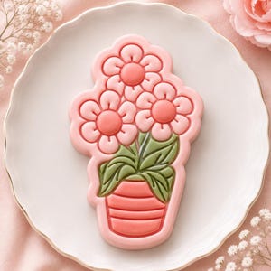 Puede incluir: Una galleta con forma de planta en maceta con tres flores rosas de centros rosa oscuro. La planta tiene hojas verdes y está en una maceta color coral con líneas horizontales. Todo el diseño está delineado por un borde rosa en relieve.