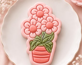 Könnte beinhalten: Ein Keks in Form einer Topfpflanze mit drei rosa Blüten und dunkelrosa Mitten. Die Pflanze hat grüne Blätter und steht in einem korallenfarbenen Topf mit horizontalen Linien. Das gesamte Design ist von einem erhabenen rosa Rand umgeben.