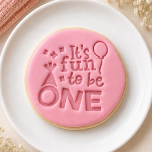 May include: A round sugar cookie topped with smooth pink fondant, featuring the embossed text It's fun to be ONE, accompanied by a party hat, a balloon, and small star shapes, centred on a plain white plate.