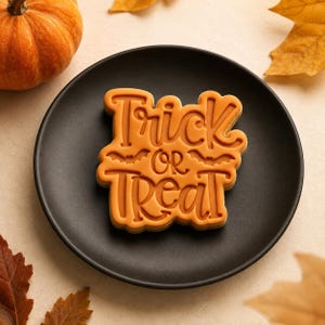 Puede incluir: Una galleta naranja con forma de placa rectangular de bordes redondeados, que presenta el texto en relieve Trick or Treat y dos pequeñas siluetas de murciélagos. La galleta está centrada en un plato negro liso, rodeada de hojas de otoño.