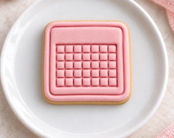 May include: A square-shaped sugar cookie decorated with smooth pink icing in the shape of a calendar. The cookie features a raised grid pattern representing the days of the month and a solid rectangular header section, all centered on a white plate.