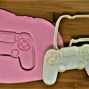 May include: A white plastic cookie cutter shaped like a video game controller. The cutter is on a wooden surface next to a pink piece of modeling clay with an impression of the cutter.
