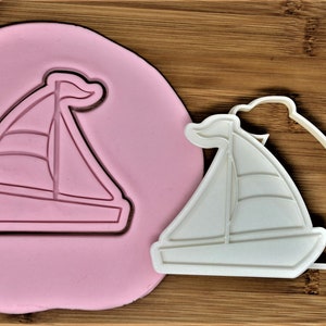 May include: A white plastic cookie cutter in the shape of a sailboat. The cutter is on a pink surface and a wooden surface.