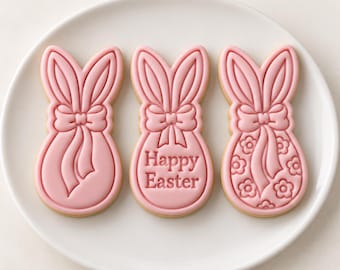 May include: Three light pink sugar cookies shaped like bunny heads with long ears and bows. The center cookie features the embossed text Happy Easter, while the right cookie displays a floral pattern. The cookies are arranged on a white plate.