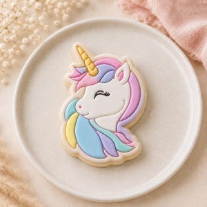 May include: A unicorn-shaped sugar biscuit decorated with white, pastel pink, blue, yellow and purple icing. The biscuit features a golden yellow horn, a closed eye with long eyelashes, and a flowing mane in multiple pastel colours on a plate.