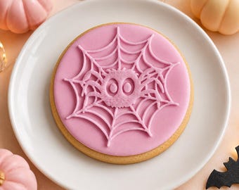 May include: A round sugar cookie decorated with smooth light pink icing featuring a raised spider web design and a central spider with two large eyes. The cookie is centered on a white plate, surrounded by small pink and cream-colored pumpkins.