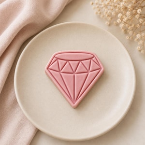 May include: A solid, matte, dusty rose-coloured wax melt shaped like a faceted diamond. The object features a geometric pattern of raised lines forming triangular and trapezoidal shapes across its surface, centred on a round, off-white ceramic plate.