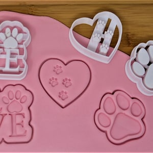 Könnte beinhalten: Ein Set aus fünf weißen Plastik-Ausstechformen in Form eines Herzens, Pfotenabdrücken und dem Wort "LOVE" mit einem Pfotenabdruck in der Mitte des Wortes. Die Ausstechformen befinden sich auf einer rosa Silikon-Backmatte.