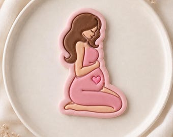 Puede incluir: Una galleta con forma de figura embarazada arrodillada con cabello largo castaño, vistiendo un vestido rosa claro. La figura tiene las manos sobre un vientre redondeado, que presenta un pequeño detalle de corazón rosa centrado. Borde rosa claro.