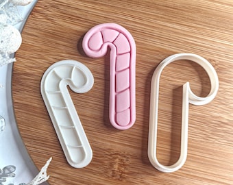 May include: Three candy cane-shaped cookie cutters. One is a solid pink colour, one is a white outline, and the other is a white cutter with interior lines. The cutters are on a wooden surface.