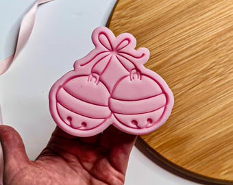 May include: A pink cookie cutter shaped like two Christmas bells joined by a bow. The bells feature horizontal lines and small circles. The cutter is held in a hand.