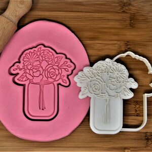 Puede incluir: Un cortador de galletas de plástico blanco en forma de tarro de cristal con un ramo de flores. El cortador de galletas está sobre una estera de hornear de silicona rosa.