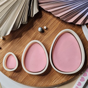 Puede incluir: Tres cortadores de galletas de plástico rosa con forma de huevo sobre una superficie de madera. Los cortadores son de diferentes tamaños, siendo el más grande el que está en el centro de la imagen.