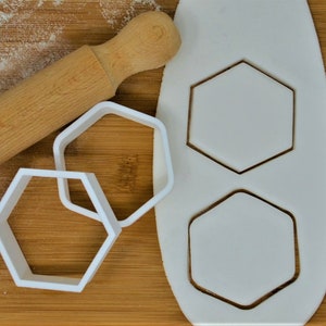 May include: White plastic cookie cutters in the shape of hexagons. The cutters are on a wooden surface with a rolling pin.