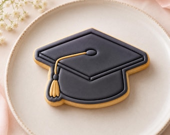 May include: A graduation cap shaped cookie decorated with smooth dark gray icing and a gold icing tassel. The cookie features a raised border and a detailed gold cord leading to a small gold tassel hanging from the side of the mortarboard.