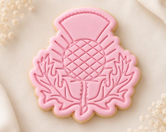 Puede incluir: Una galleta con forma de flor de cardo, con una capa de glaseado rosa claro liso y un patrón geométrico grabado que representa la cabeza de la flor y hojas espinosas, sobre un fondo liso de color claro.