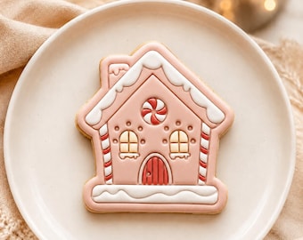 Puede incluir: Una galleta con forma de casa decorada con glaseado rosa, detalles de glaseado blanco en el techo y la base, dos ventanas arqueadas con cristales amarillos, una puerta arqueada roja y un detalle de caramelo de menta rojo y blanco.