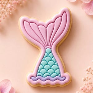 May include: A mermaid tail shaped sugar biscuit decorated with light pink icing on the upper fin and teal blue icing on the lower section featuring a raised scale pattern, finished with a thin purple wavy line at the base of the tail design.