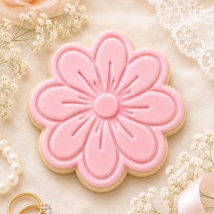 May include: A flower-shaped sugar biscuit decorated with smooth, light pink royal icing. The biscuit features a central circular detail surrounded by eight rounded petals, each defined by embossed lines that create a clean, raised texture.
