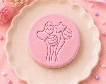 May include: A round, light pink cookie featuring an embossed design of a cluster of balloons in various shapes, including a heart, a star, and round balloons with striped and wavy patterns, centered on a scalloped-edge light pink plate.