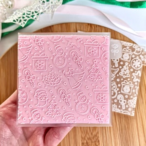 Puede incluir: Carpeta de relieve cuadrada rosa con un patrón navideño. El diseño incluye un muñeco de nieve, un árbol de Navidad, una caja de regalo, un bastón de caramelo, un ángel y Papá Noel. Una pestaña de plástico transparente está adjunta.