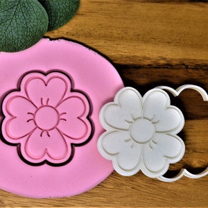 Puede incluir: Un cortador de galletas de plástico blanco en forma de flor con una impresión de fondant rosa del mismo diseño de flor.