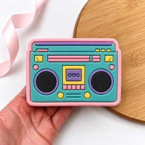 May include: A rectangular boombox-shaped object featuring a teal main body with a pink border. It includes two black circular speakers, a central yellow cassette deck with a purple cassette, and various yellow and pink buttons and dials.