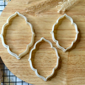 May include: Three white plastic cookie cutters in the shape of an ornate oval frame. The cutters are on a wooden surface.