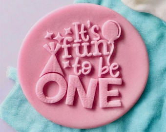 May include: A pink, circular fondant cake topper with the phrase "It's fun to be ONE" embossed in white. The design includes a party hat, stars, and a balloon. The topper is set on a blue and white fabric background.