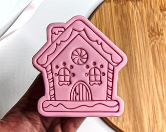 May include: A pink gingerbread house cookie cutter. The cutter features a detailed design with a chimney, windows, and a door. The roof has a scalloped edge, and the house has a candy swirl detail. This baking tool is perfect for creating festive biscuits.
