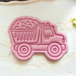 May include: A pink plastic cookie cutter shaped like a dump truck. The truck bed is filled with a detailed design of rocks. The wheels and cab are also detailed. The cookie cutter is on a white surface.