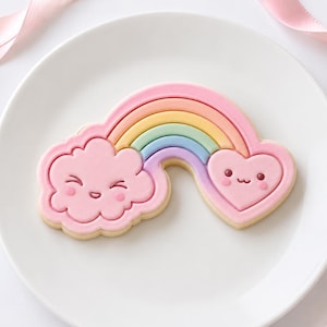 May include: A sugar cookie shaped like a rainbow connecting a pink cloud and a pink heart. The cloud and heart both feature simple, hand-drawn style faces with eyes and a mouth. The rainbow consists of pastel yellow, green, blue, and purple stripes.