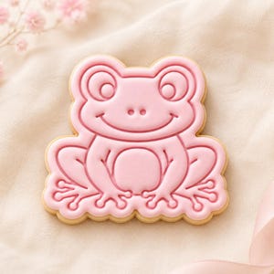 May include: A light pink sugar biscuit shaped like a frog with embossed facial features, large eyes, and webbed feet. The biscuit has a smooth, matte icing finish and a light tan edge, showcasing a detailed outline of the frog's body and limbs.
