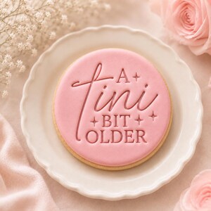 May include: A round sugar cookie with smooth pink icing featuring the embossed text A Tini BIT OLDER and small star accents. The cookie is centered on a white scalloped plate, showcasing a clean, flat surface with clear, legible lettering.