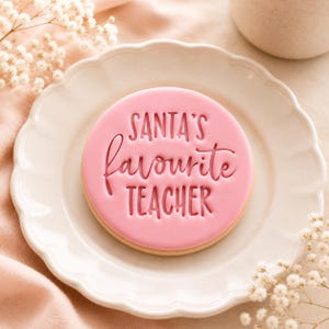 May include: A round sugar cookie topped with smooth pink icing. The text SANTA'S favourite TEACHER is embossed into the center of the icing in a combination of bold block lettering and a cursive script font, creating a clear, readable message.
