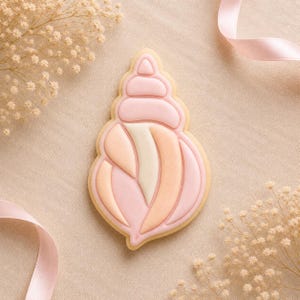 May include: A seashell-shaped biscuit decorated with smooth icing in shades of soft pink, peach, and cream. The design features a raised, segmented top section and a lower body with curved, vertical panels, all outlined with fine piping.