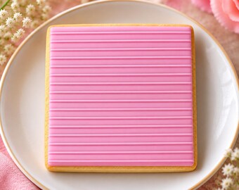 Puede incluir: Una galleta cuadrada cubierta con una capa de glaseado rosa brillante y suave, que presenta una serie de líneas horizontales en relieve espaciadas uniformemente, creando un patrón de rayas texturizado en la superficie.