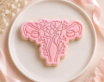 May include: A light pink cookie shaped like a human uterus, featuring an embossed floral design with leaves and petals across the surface. The cookie is centered on a plain white plate, showcasing detailed line work and a smooth, matte finish.