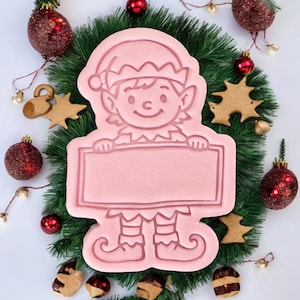 Puede incluir: Un cortador de galletas rosa con forma de elfo con un letrero en blanco, rodeado de decoraciones navideñas. El cortador presenta un elfo con sombrero y botas. El fondo incluye una corona verde, adornos rojos y galletas en forma de estrella.