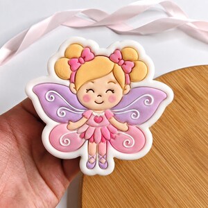 May include: A sugar cookie shaped like a fairy with blonde hair in two buns, pink bows, and a pink dress with a heart detail. The fairy has purple and pink wings with white swirl patterns, rosy cheeks, and purple ballet slippers on its feet.