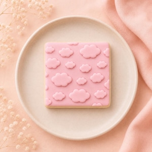 May include: A square biscuit with a smooth, light pink icing base featuring a repeating pattern of raised, slightly darker pink clouds. The biscuit is centred on a round, off-white ceramic plate, showcasing a clean and uniform design.